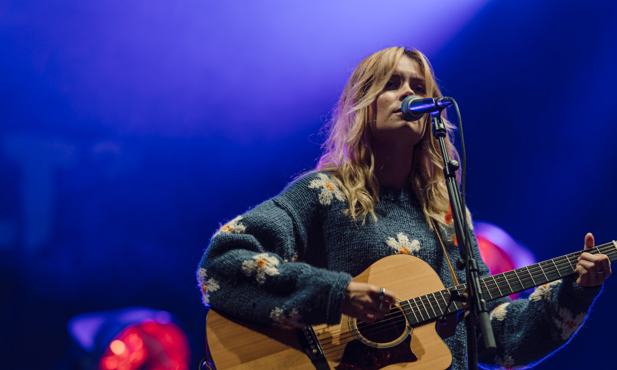 Nina Nesbitt plays guitar while wearing a flower pattern jumper