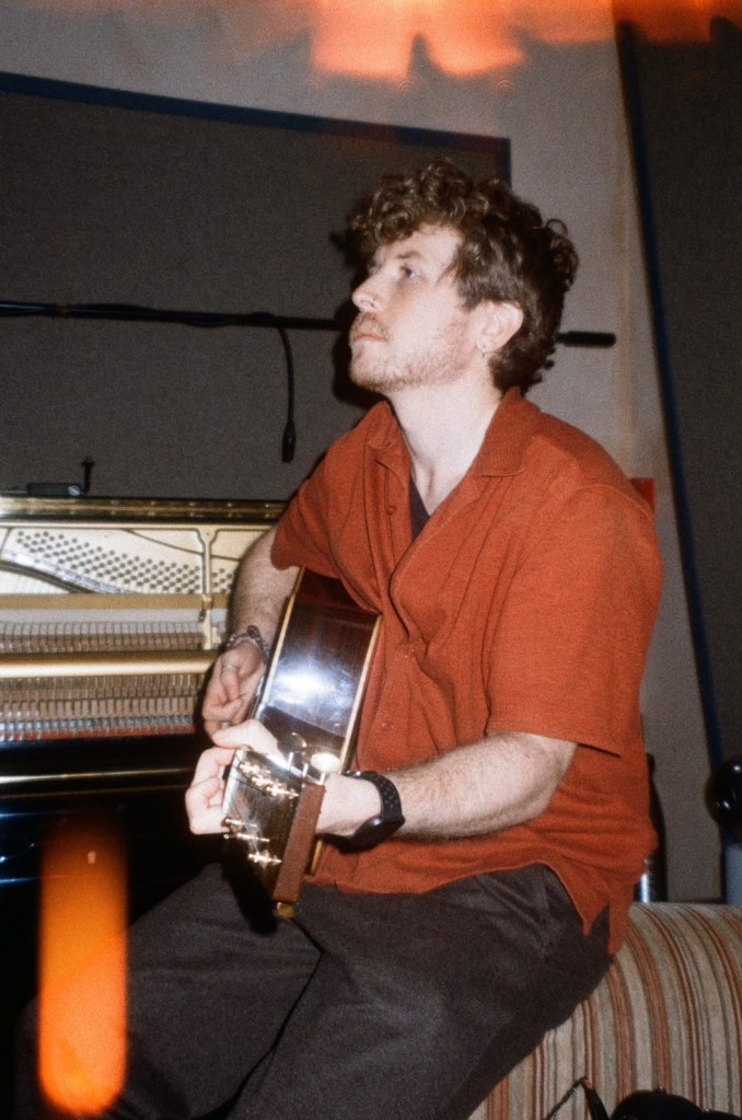 A man with curly hair sits on a couch, playing an acoustic guitar in a recording studio, with a piano visible in the background.