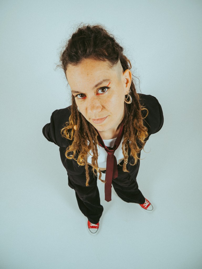 A person with dreadlocks and a stylish outfit, including a black blazer, white shirt, and red tie, standing with a confident expression against a light blue background.
